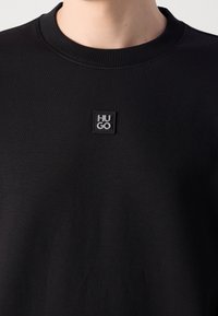 T-shirt noir à col rond avec un petit patch carré noir sur la poitrine affichant les lettres blanches "HU GO".