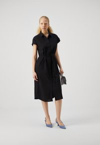 Rochie neagră cu guler, cu un cordon în talie, mâneci scurte și o țesătură texturată. Asortată cu pantofi slingback bleu deschis și o mică geantă gri.