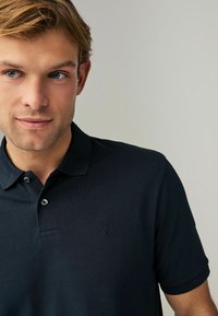 Mørkeblå polo-shirt lavet af tekstureret bomuld. Den har en klassisk krave, en to-knaps placket og et lille logo broderet på brystet.