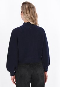 Navyblauwe geribbelde trui met een hoge kraag en lange pofmouwen. Heeft een aansluitende tailleband en een klein logo-detail aan de achterkant.