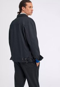 Giacca nera in misto lana con colletto strutturato, dettagli dei bottoni e tasche laterali. Il modello indossa una camicia blu con i polsini risvoltati sotto.