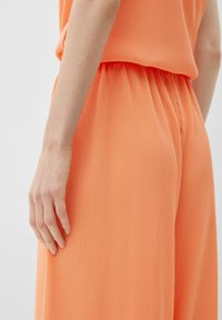 Broek gemaakt van ribbelige, textuurstof in feloranje. Elastische tailleband en een losse pasvorm, met verticale strepen. Hand rustend op de stof.