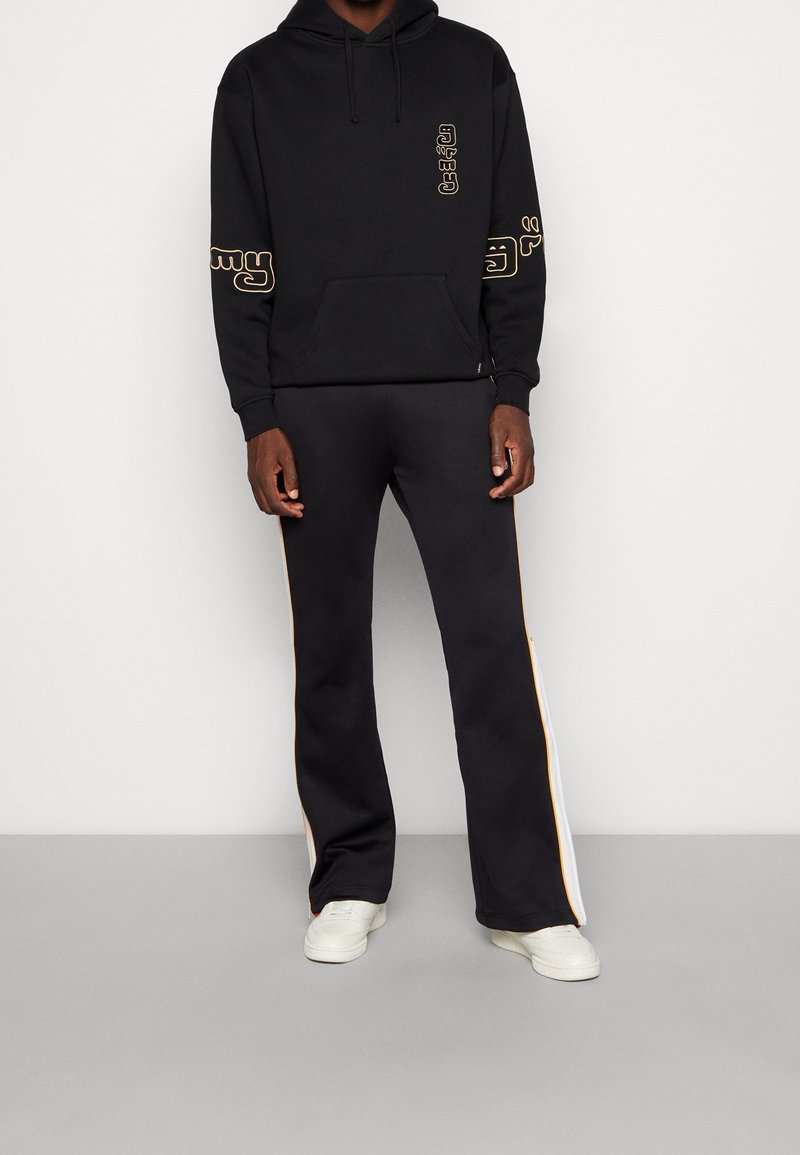 Homme portant un sweat à capuche noir avec un texte graphique jaune sur les manches et un pantalon noir assorti avec des bandes latérales jaunes et blanches, baskets blanches.