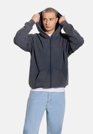 Ciemnoniebieska bluza z kapturem na zamek wykonana z miękkiego materiału, z przednią kieszenią, kapturem ze sznurkiem i wyszywanym logo na piersi. Połączona z jasnymi jeansami.