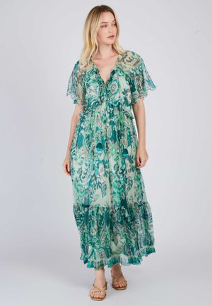 Woman wearing a flowing green and teal floral maxi dress with ruffled short sleeves and V-neck, standing against a plain light background.