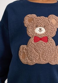 Granatowy sweatshirt z teksturowanym wzorem brązowego misia teddy z czerwoną muszką, wykonany z miękkiej, pluszowej tkaniny i wyraźnymi szczegółami szycia.