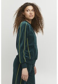 Felpa in velluto di colore verde scuro con strisce gialle ai lati, scollo rotondo, tasca frontale e polsini a coste sulle maniche e sull'orlo.