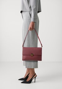 Μπορντό δερμάτινη τσάντα με υφασμάτινη υφή, διαθέτει κούμπωμα από χρυσό υλικό και αποσπώμενο λουρί, την κρατάει ένα άτομο με καρό παντελόνι.