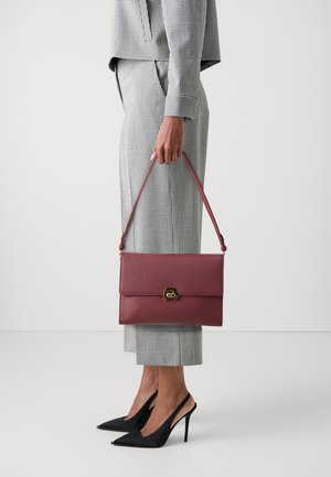 Borsa a mano in pelle bordeaux con finitura texture, dotata di chiusura in metallo dorato e tracolla removibile, tenuta da una persona con pantaloni quadrettati.