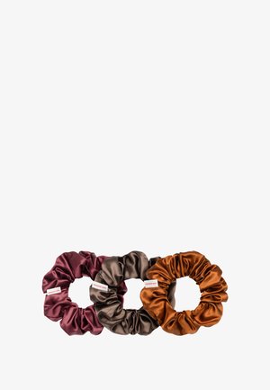 Trois chouchous en satin de couleur bordeaux, marron et rouille. Chacun présente une texture lisse avec un tissu froncé et une petite étiquette de marque.