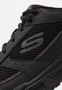 Schwarzer Sportschuh mit Netzpaneelen, strukturiertem synthetischem Material, abgerundeter Spitze, Schnürdesign und Gummisohle mit Profil.