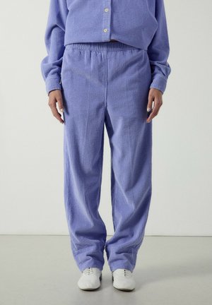 Person trägt locker sitzende lavendelfarbene Cordhose mit elastischem Bund und passendem Hemd mit Knöpfen, kombiniert mit weißen Schnürschuhen.