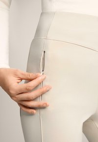 Beżowe legginsy z wysokim stanem mają zasuwaną kieszeń po boku. Materiał jest gładki z subtelnymi detalami szwów. Ręka pokazana w interakcji z zamkiem błyskawicznym.
