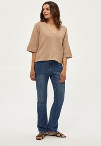 Beige V-hals sweater med korte ærmer, parret med mørkeblå jeans og sorte sandaler med gyldne nitter. Casual outfit præsenteret.