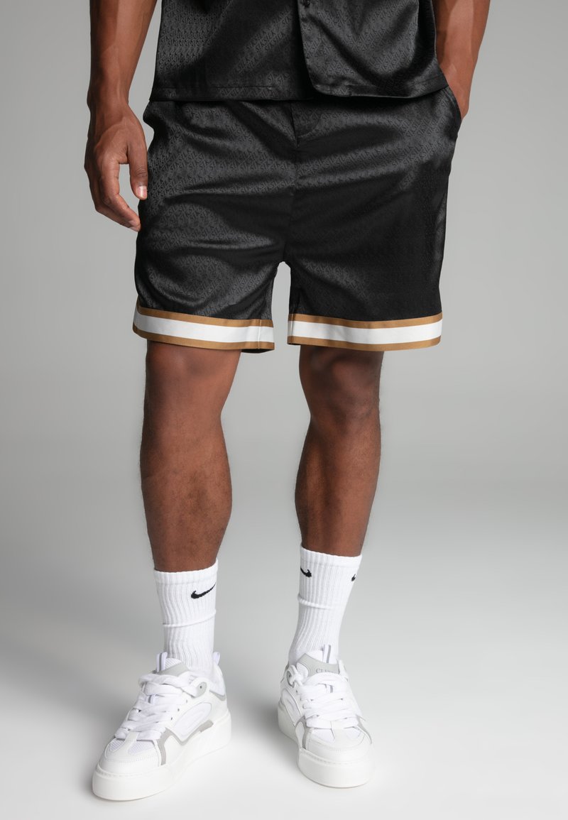 Shorts en satin noir avec un motif subtil, ourlet rayé doré et blanc, associés à des baskets blanches et des chaussettes Nike. Texture lisse.