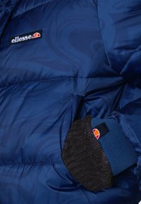 Niebieska, pikowana kurtka z wirującą teksturą, z czarnym dzianinowym mankietem i logo ellesse na klatce piersiowej. Kurtka o średniej długości.