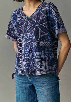 Femme portant une chemise bleue à manches courtes col en V avec des motifs patchwork associée à un jean bleu sur fond uni.