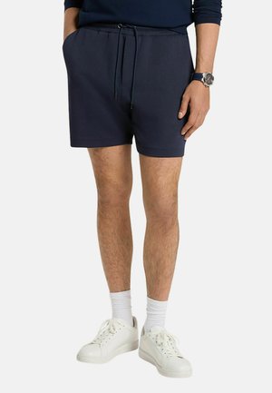 Uomo che indossa pantaloncini blu navy con coulisse, calzini bianchi, scarpe da ginnastica bianche e un orologio da polso, in piedi con una mano in tasca.