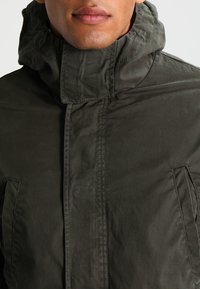 Giacca verde scuro con colletto alto, dotata di chiusura con zip e due tasche laterali. Realizzata in un materiale leggero e liscio con una finitura opaca.