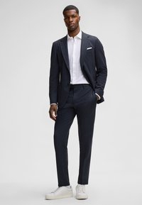 Dunkel gemustertes Anzug mit taillierter Silhouette, ausgestattet mit einem Reverskragen, weißem Hemd darunter und hellen Sneakern.