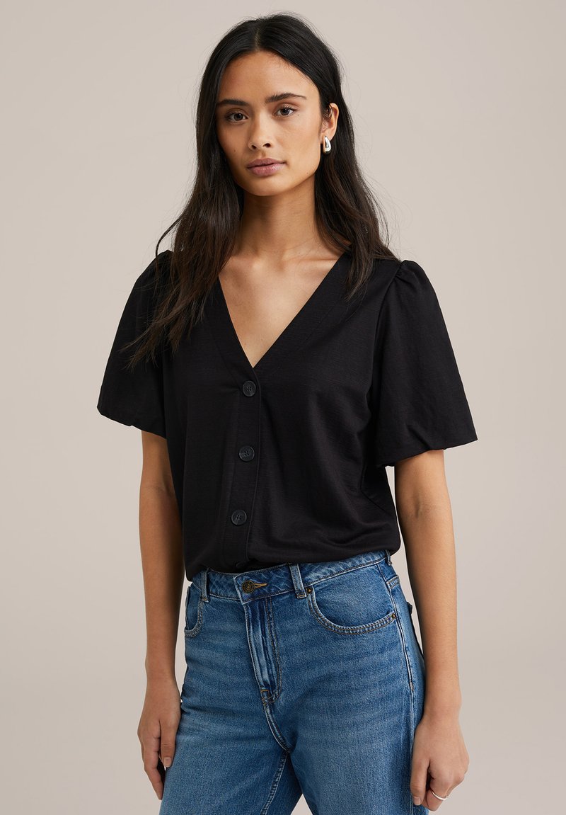 Črn kratkosleeven top z V-izrezom in štirimi gumbi spredaj; gladka tekstura; nošene z modrimi hlačami na visoko pasu.