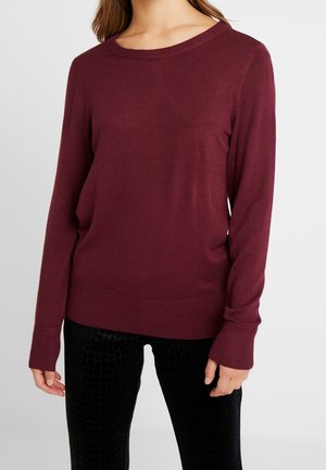 Maglione a maglia bordeaux con scollatura rotonda e maniche lunghe, caratterizzato da polsini e orlo a coste, abbinato a pantaloni neri con texture.