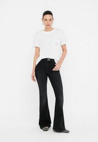 T-shirt bianco a maniche corte con taschino sul petto; abbinato a jeans neri a palazzo. L'outfit è completato da una cintura nera e scarpe con fantasia.