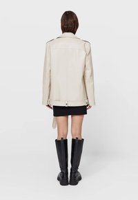 Cremfarbene Lederjacke mit langen Ärmeln, strukturierten Schultern und einem taillierten belted Design, kombiniert mit einem schwarzen Minirock und knielangen schwarzen Stiefeln.