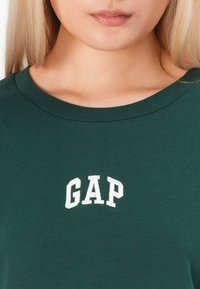 Skoggrønn t-skjorte med rund hals og hvit brodert "GAP"-logo sentrert på brystet. Glatt, bomullstoff.
