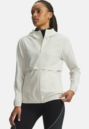 Frau trägt eine weiße Kapuzenjacke mit Reißverschluss und schwarze Leggings mit gemusterten Seitenteilen, steht mit einer Hand auf der Hüfte.