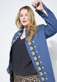 Modrá košile na knoflíky s vyšívanými detaily na přední straně, s límečkem a uvolněnými rukávy, přes texturovaný černý top a vzorované kalhoty.