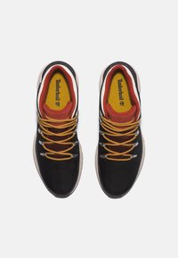 Mustat verkkomaiset lenkkarit, joissa on keltaisia ja oransseja yksityiskohtia, paksut mustat nauhat, kumipohja ja Timberland-logo näkyvissä sisällä.