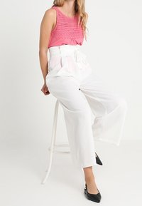 Lyserød ærmeløs top med tekstureret syning, båret sammen med højtaljede hvide bredbenede bukser og spidse sorte hæle, siddende på en skammel.