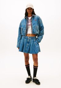 Chaqueta vaquera de diseño corto, falda vaquera azul plisada, camisa blanca con texto, gorra beige y calcetines negros con rayas rojas y blancas.