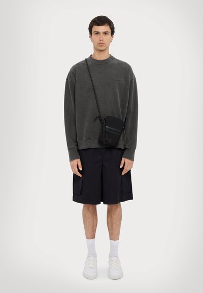 Grauer Sweatshirt mit Rundhalsausschnitt, lockerer Passform, kombiniert mit schwarzen Shorts und weißen Sneakers. Mit einer kleinen schwarzen Umhängetasche.