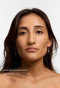 Base de maquillaje de tono cálido ligero a medio, etiquetada con el nombre del producto. La modelo tiene el cabello largo, lleva pendientes de espina de pescado y un choker rosa delgado, con fondo neutro.