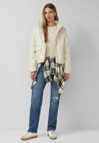 Pikowana kremowa kurtka z przednimi kieszeniami, noszona na beżowej bluzce. Jeansy o luźnym kroju i koszula w kratę związana w pasie, w połączeniu z białymi trampkami.