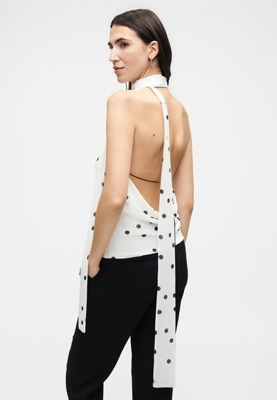 Femme aux longs cheveux foncés portant un haut blanc à pois avec un dos ouvert et un pantalon noir, posant les mains dans les poches.