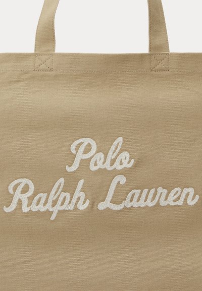 Μπεζ τσάντα ώμου από καμβά με δύο λαβές και κεντημένο λευκό κείμενο στην πρόσοψη που αναγράφει "Polo Ralph Lauren".