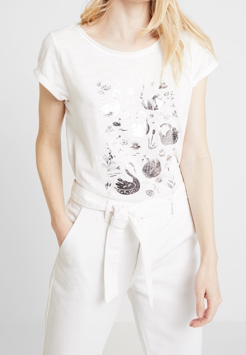 Camiseta blanca de manga corta con un gráfico de cisne en tono plateado y flores. Combinada con pantalones blancos de tiro alto y un cinturón anudado.