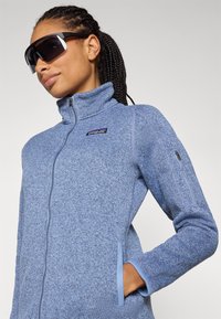 Giacca in pile blu con zip, colletto strutturato, tasche laterali e patch del logo. La texture è morbida, con accenti di un blu chiaro contrastante.