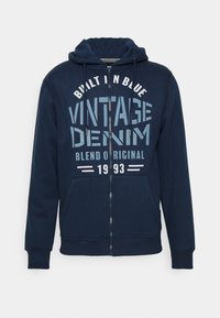 Mornarska zip-up jopica iz bombažne mešanice, z grafičnim tiskom in napisom "VINTAGE DENIM" ter sprednjimi žepi. Nastavljiv kapuco z vrvico.