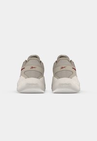 Zapatillas atléticas beige con parte superior de malla texturizada y suela de goma. Presenta un sutil acento rojo cerca del talón y el logotipo de la marca en la parte posterior.