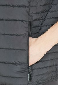 Schwarze gepolsterte Weste mit horizontalen Steppnähten, ausgestattet mit einer seitlichen Reißverschlusstasche und einem Logo-Aufnäher. Der Stoff wirkt glatt und leicht.