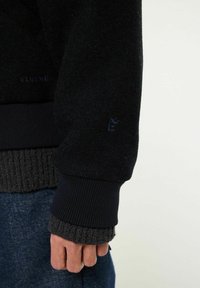 Svart stickad tröja med ribbad nederkant, försedd med en texturerad manschett. Diskret logotyp på ärmen. Materialet verkar mjukt och texturerat.