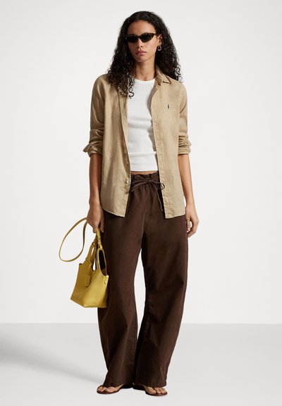 Kobieta w okularach przeciwsłonecznych w szkielecie w kolorze żółwia, beżowej koszuli zapinanej na guziki na białą bluzkę, brązowych spodniach o szerokich nogawkach, trzymająca żółtą torebkę, stojąca na białym tle.