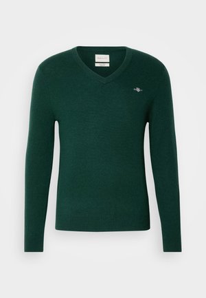 Dunkelgrüner V-Ausschnitt-Pullover aus weichem Material. Figurbetonter Schnitt mit langen Ärmeln und einem kleinen bestickten Logo auf der Brust.
