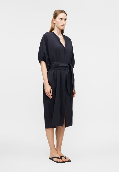 Γυναίκα που στέκεται φορώντας ένα σκούρο μπλε φόρεμα μέχρι το γόνατο με ζώνη δεμένη στη μέση και μαύρα σαγιονάρες σε απλό φωτεινό φόντο.