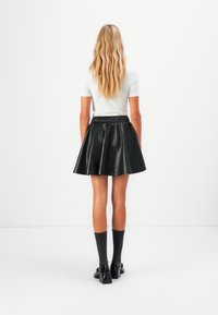 Žena s dlouhými vlnitými blond vlasy, oblečená v bílém topu s krátkým rukávem, černé kožené sukni, šedých ponožkách a černých lodičkách, stojí čelem odvrácena.
