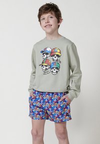 Sudadera verde claro con calaveras en gorros coloridos, combinada con pantalones cortos azules que presentan un patrón de calaveras rojas, amarillas y azules.